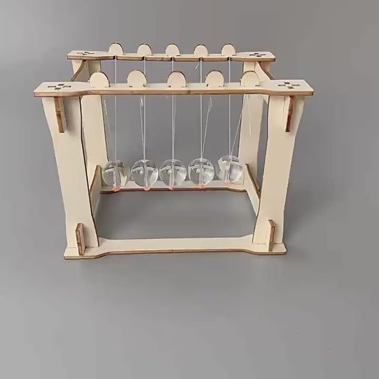 DIY Newton's Cradle Kit assembly steps shown. Wooden frame, acrylic spheres, and string components being put together. Educational STEM toy.