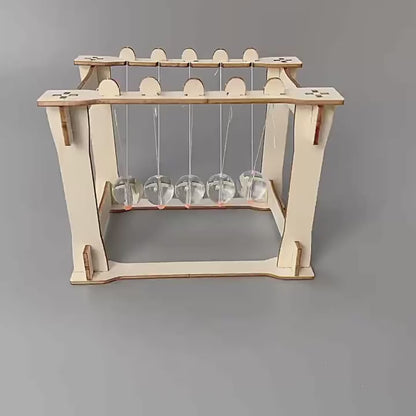 DIY Newton's Cradle Kit assembly steps shown. Wooden frame, acrylic spheres, and string components being put together. Educational STEM toy.